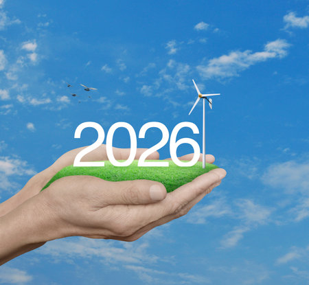 2026 white text with wind turbine on green grass field in hands over blue sky, white clouds and...