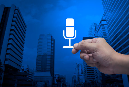 Hand holding microphone flat icon in fingers over modern office city tower and skyscraper, Technology communication conceptの写真素材