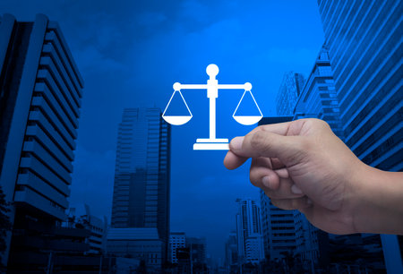 Hand holding law flat icon in fingers over modern office city tower and skyscraper, Business legal service conceptの写真素材