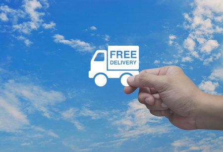 Hand holding free delivery truck flat icon in fingers over blue sky with white clouds, Business transportation service conceptの写真素材