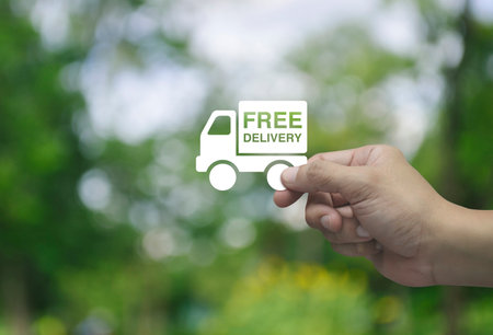 Hand holding free delivery truck flat icon in fingers over blur green tree in park, Business transportation service conceptの写真素材