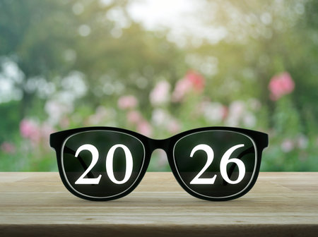 2026 white text with black eye glasses on wooden table over blur pink flower and tree in garden,...