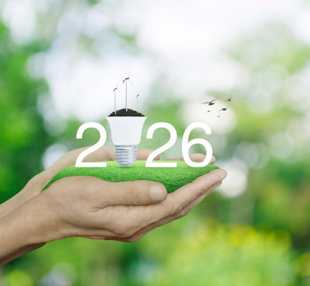 2026 white text with wind turbines on soil and light bulb on green grass in hands over blur forest, Happy new year 2026 green ecology and saving energy conceptの写真素材