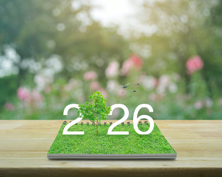 2026 white text with growing tree on green grass on open book on wooden table over blur pink flower and tree in garden, Happy new year 2026 ecological cover conceptの写真素材