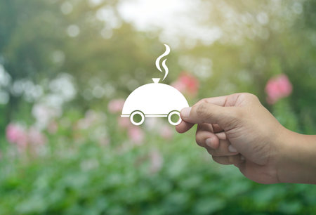 Hand holding restaurant cloche flat icon in fingers over blur pink flower and tree in park, Food delivery online service conceptの写真素材