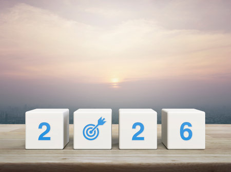 2026 letter with goal flat icon on white block cubes on wooden table over modern city tower and...