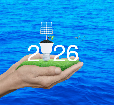 2026 white text with solar cell and fresh leaves on soil and light bulb on green grass in hands over...
