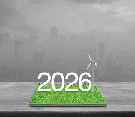 2026 white text with wind turbine on green grass on open book on wooden table over pollution city...