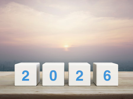 2026 number on white block cubes on wooden table over modern city tower and skyscraper at sunset,...