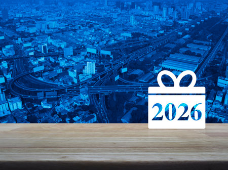 Gift box happy new year 2026 flat icon on wooden table over modern city tower, street, expressway...