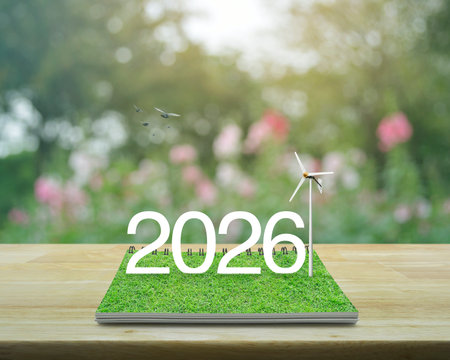 2026 white text with wind turbine on green grass on open book on wooden table over blur pink flower...