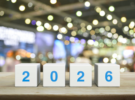 2026 letter on white block cubes on wooden table over blur light and shadow of shopping mall, Happy...
