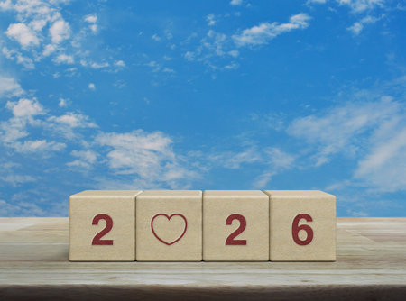 Love flat icon with 2026 letter on wood block cubes on wooden table over blue sky with white clouds,...