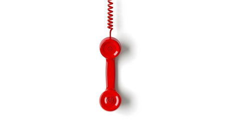 Vintage red telephone handset suspended by spiral cord on plain white background. Retro communication device, obsolete technology symbol. Object isolated with shadow.の素材