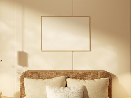 Small square frame with blank poster above soft bed pillows in a warm room.の写真素材
