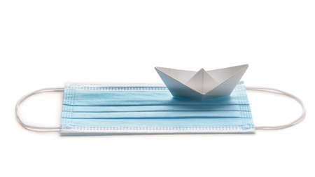 Sailing during pandemic crisis. Surgical mask with white paper ship on white background.の写真素材