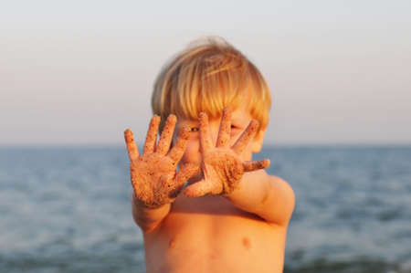 Little boy plays with sand on the beach at evening sunset, showing his handsの写真素材