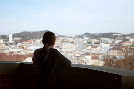 Rear view of lonely eleven years girl with long blond hair standing on balcony and looking to panorama of Lviv in spring,の写真素材