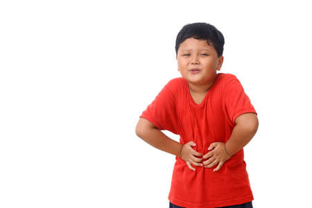 Close up photo of asian boy in red shirt standing while holds his stomach, he feels pain. Isolated on white background with copy spaceの写真素材