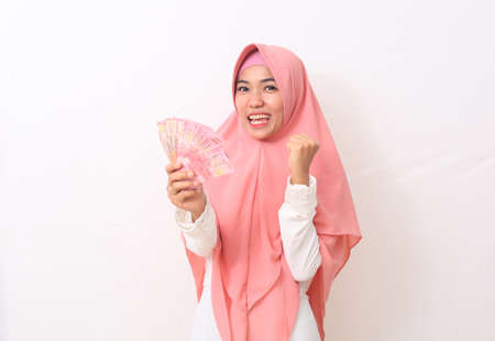 Happy asian woman in Muslim dress stands up, holding Indonesian money. Isolated on white backgroundの写真素材