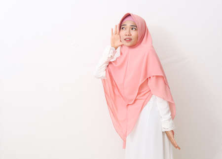 A portrait of happy asian muslim woman wearing a veil or hijab smiling and doing hearing gesture, put hand on ear, listening carefully. Isolated on white background with copy spaceの写真素材