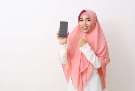 A portrait of happy asian muslim woman wearing a pink veil or hijab and white dress showing and pointing a blank smartphone screen. White background with copy spaceの写真素材