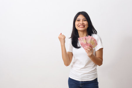 Happy asian girl holds an Indonesian money while standing, looking at camera and clenched her fists....