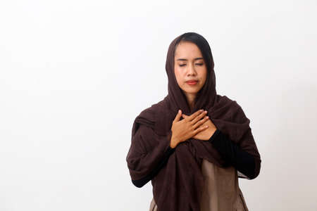 Faithful asian muslim woman closes eyes and keeps hands on chest near heart, shows kindness or favour, expresses sincere emotions, being kind hearted and honest. Body language, real feelings conceptの写真素材