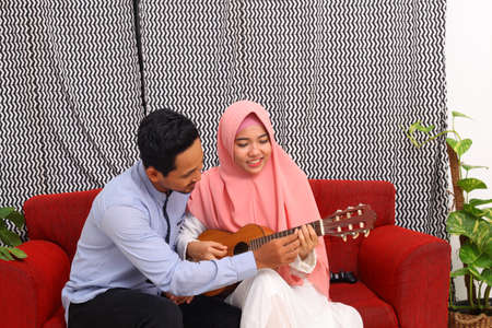 Asian muslim couple sitting on red sofa while learn to playing guitar