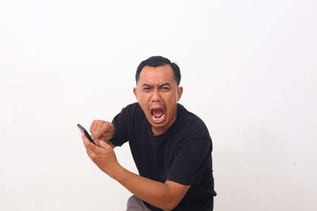 Attractive young Asian man pointing on his phone, getting bad news, shocked mad angry gesture over whiteの写真素材