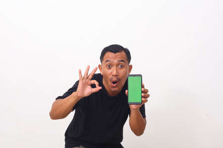 Portrait of satisfied asian man with beard in black t-shirt pointing at cellphone and smiling at camera, recommending gadget or mobile application. indoor studio shot isolated on white backgroundの写真素材