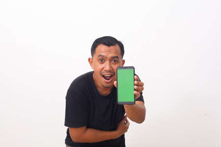 Portrait of satisfied asian man with beard in black t-shirt pointing at cellphone and smiling at...