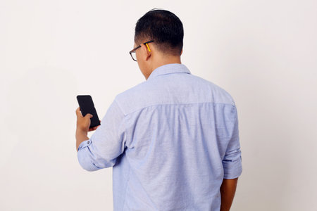 Back view of Adult Asian man wearing bright blue shirt looking to mobile phone screen. Isolated on whiteの写真素材
