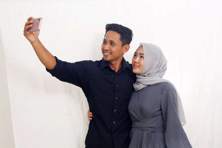 Joyful asian muslim couple making selfie using their cellular phone. Isolated on whiteの写真素材