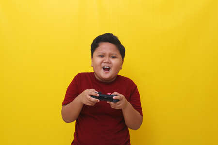 Rousing asian boy standing while holding a joystick. Gaming concept. Isolated on yellow backgroundの写真素材
