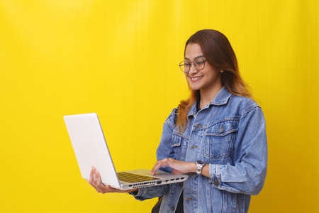 Happy asian young college student standing while holding a laptop. Isolated on yellow backgroundの写真素材