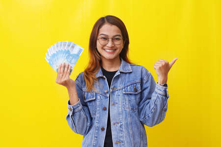 Pretty young asian woman standing while holding an Indonesian money and pointing something.の写真素材