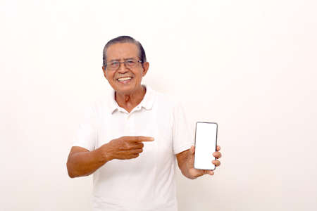 Elderly asian man standing while pointing at blank cell phone screen. Isolated on white backgroundの写真素材