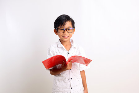 Smart asian schoolboy carrying a book while looking at the camera. Isolated on white background