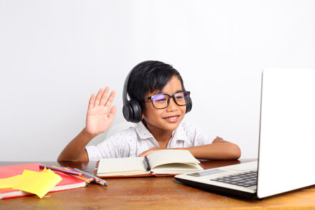 Happy asian elementary schoolboy raising hand while studying online using laptop at homeの写真素材