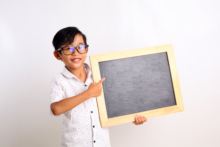 Happy asian schoolboy standing while showing a blank blackboard. Isolated on white background