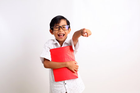 Adorable asian schoolboy carrying a book while laughing and pointing forward. Isolated on whiteの写真素材