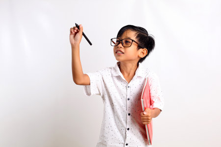 Smart asian boy standing while writing something and carrying a book. Isolated on white background