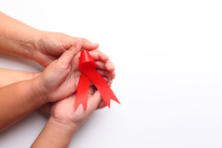 Woman's hand and child's hand holding red ribbon together over white with copyspace. Aids day...