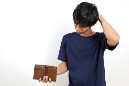 Disappointed young asian man standing while opening his empty wallet. Isolated on white backgroundの写真素材