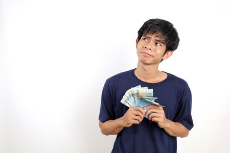 Young asian man standing while holding Indonesian banknotes and imaging something. Isolated on whiteの写真素材