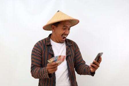 Surprised asian farmer standing while holding Indonesian banknotes and cell phone. Isolated on white...