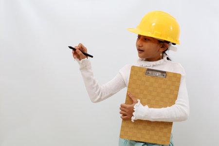 Happy Asian little girl in the construction helmet as an engineer standing while writing something....