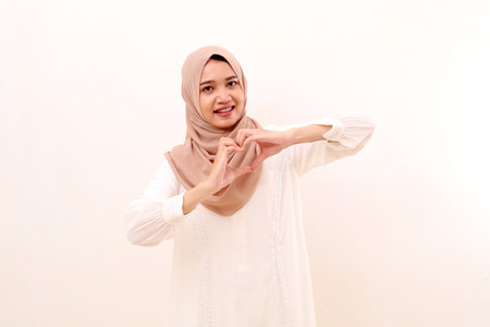 Cheerful young asian muslim woman making love heart shape with her hands. Isolated on white