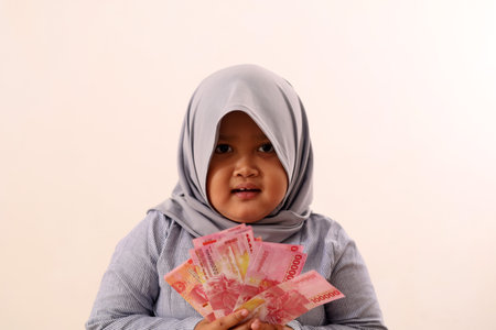 Happy asian muslim little girl holding indonesian banknotes. Isolated on white backgroundの写真素材